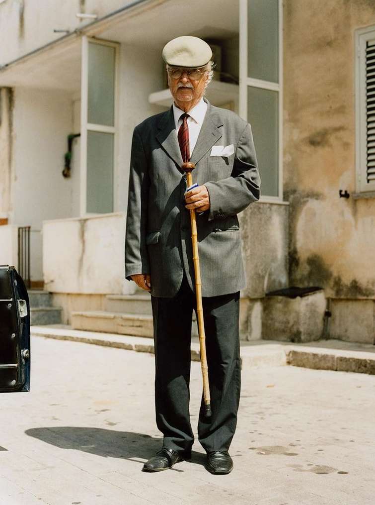 Mature man in a grey suit and cap stands holding a cane outside a building with white trim, under bright daylight.