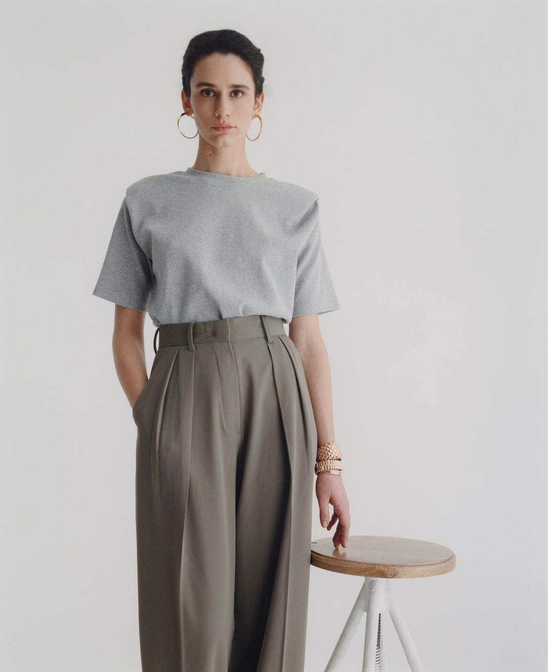 A stylish woman in a heather grey tee shirt and olive green slacks poses in a studio. She stands next to a wooden stool.