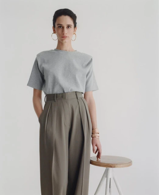 A stylish woman in a heather grey tee shirt and olive green slacks poses in a studio. She stands next to a wooden stool.