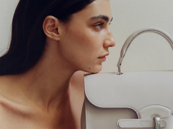 Profile portrait of a woman resting her chin on a light grey leather handbag against a plain background