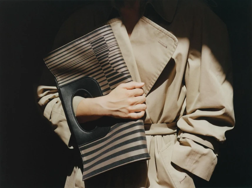 Person holds a striped clutch with a handle, wearing a tan trench coat, captured in a fashion-forward, high-contrast studio setting.