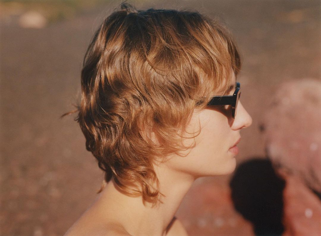 Profile portrait of a young adult woman with blonde wavy hair, wearing sunglasses, with blurry background and warm lighting.