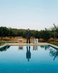 Man standing by a swimming pool with two patterned chairs flanked by trees under a clear sky, photographed by Carlo Piro.