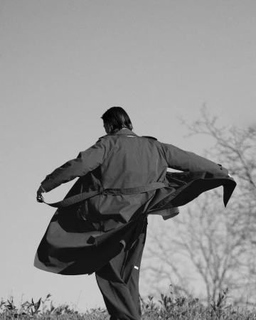 Monochrome fashion photograph of a person from behind wearing a long coat with the fabric billowing outward in the wind.