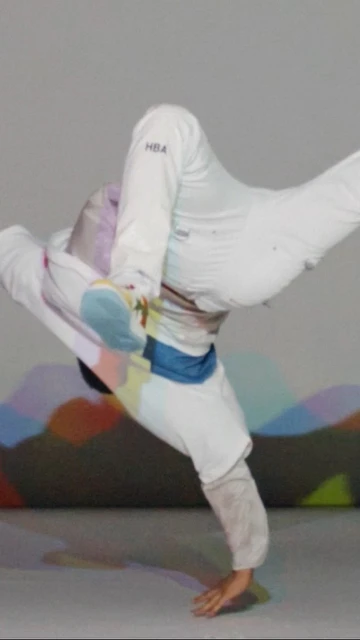 Person in dynamic breakdance pose wearing white sportswear, light grey background with abstract mountain-like shapes.