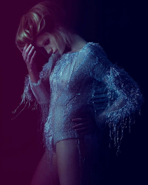 Blonde model in a shimmering silver bodysuit poses in a dark studio with dramatic lighting, highlighting the texture and form of the garment.