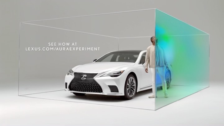White Lexus sedan in a transparent display case with a man standing near a translucent, colorfully lit panel in a neutral studio setting.