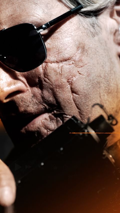 Close-up portrait of a man wearing sunglasses, featuring scars on his face, with dramatic lighting and a shallow depth of field.