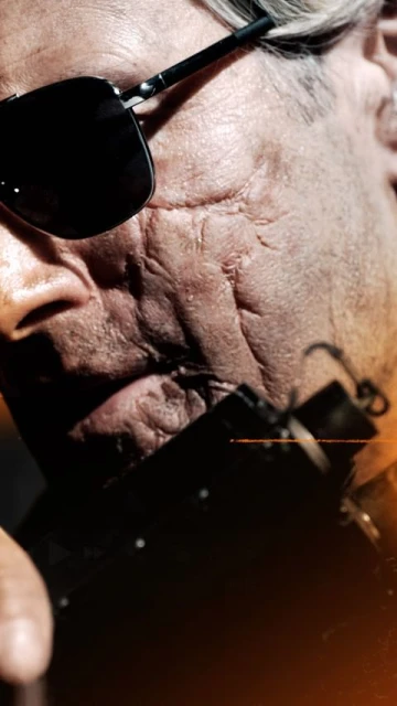 Close-up portrait of a man wearing sunglasses, featuring scars on his face, with dramatic lighting and a shallow depth of field.