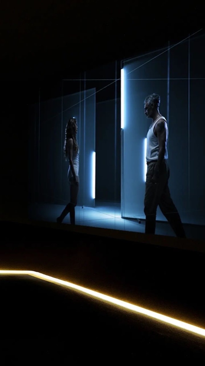 Two adults in a dimly lit indoor setting with geometric lights; an adult male and female face each other in a minimalist, contemporary scene.