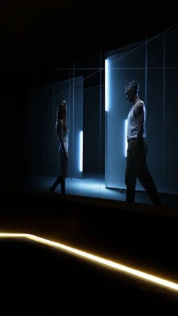 Two adults in a dimly lit indoor setting with geometric lights; an adult male and female face each other in a minimalist, contemporary scene.
