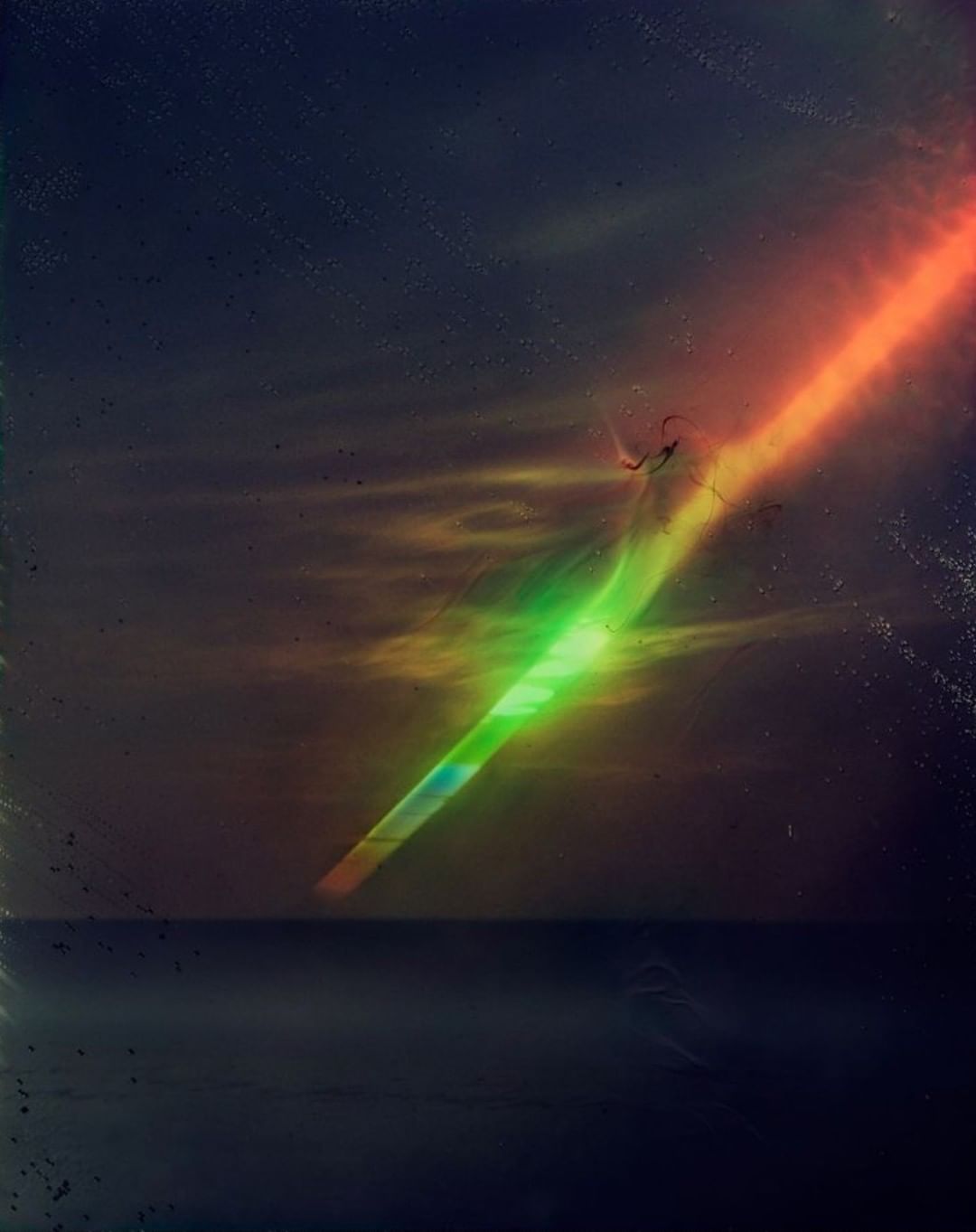 Abstract image of a colorful light beam stretching across a dark sky over water, with an orange and green glow.