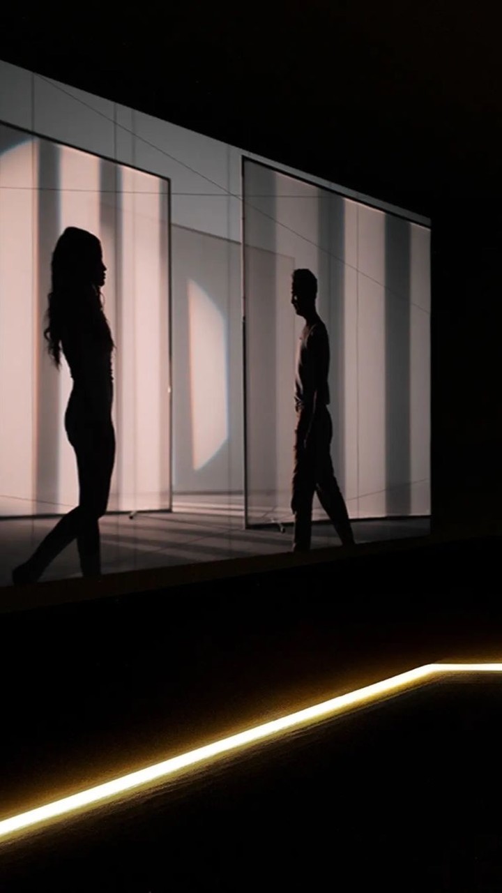 Silhouette of an adult woman and man in a dark setting, illuminated by light panels in the background. Low-angle shot.