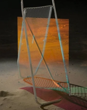 A metal-framed chair sits on a sandy surface in front of an orange-toned backdrop, casting colorful shadows with turquoise and pink hues.