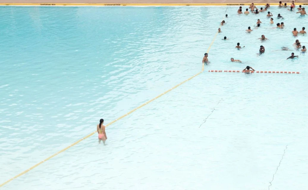 Aerial view of a large swimming pool with turquoise water, people swimming and wading, separated by a yellow rope in sunlight.