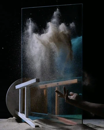 Abstract image of dust suspended in the air after being thrown at a sheet of glass, with a hand and a chair also visible.