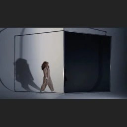 Woman in beige jumpsuit walking past a large metallic frame partition in a studio setting