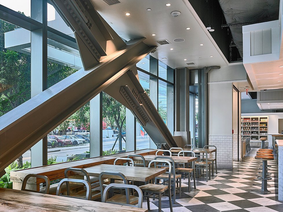 Interior view of a restaurant with metal support beams, tables and chairs, a black and white checkered floor, and large glass windows.