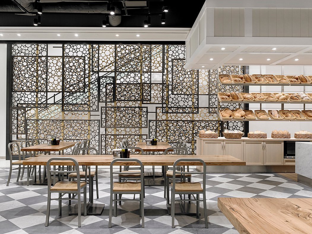 Interior view of a bakery cafe with tables and chairs on a tiled floor, and bread displayed on shelves in baskets against a wall.