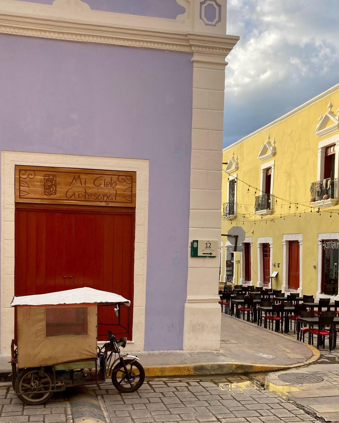 View of a vibrant yellow building with red door and iron balconies, contrasted by a periwinkle building and a bicycle taxi in front.