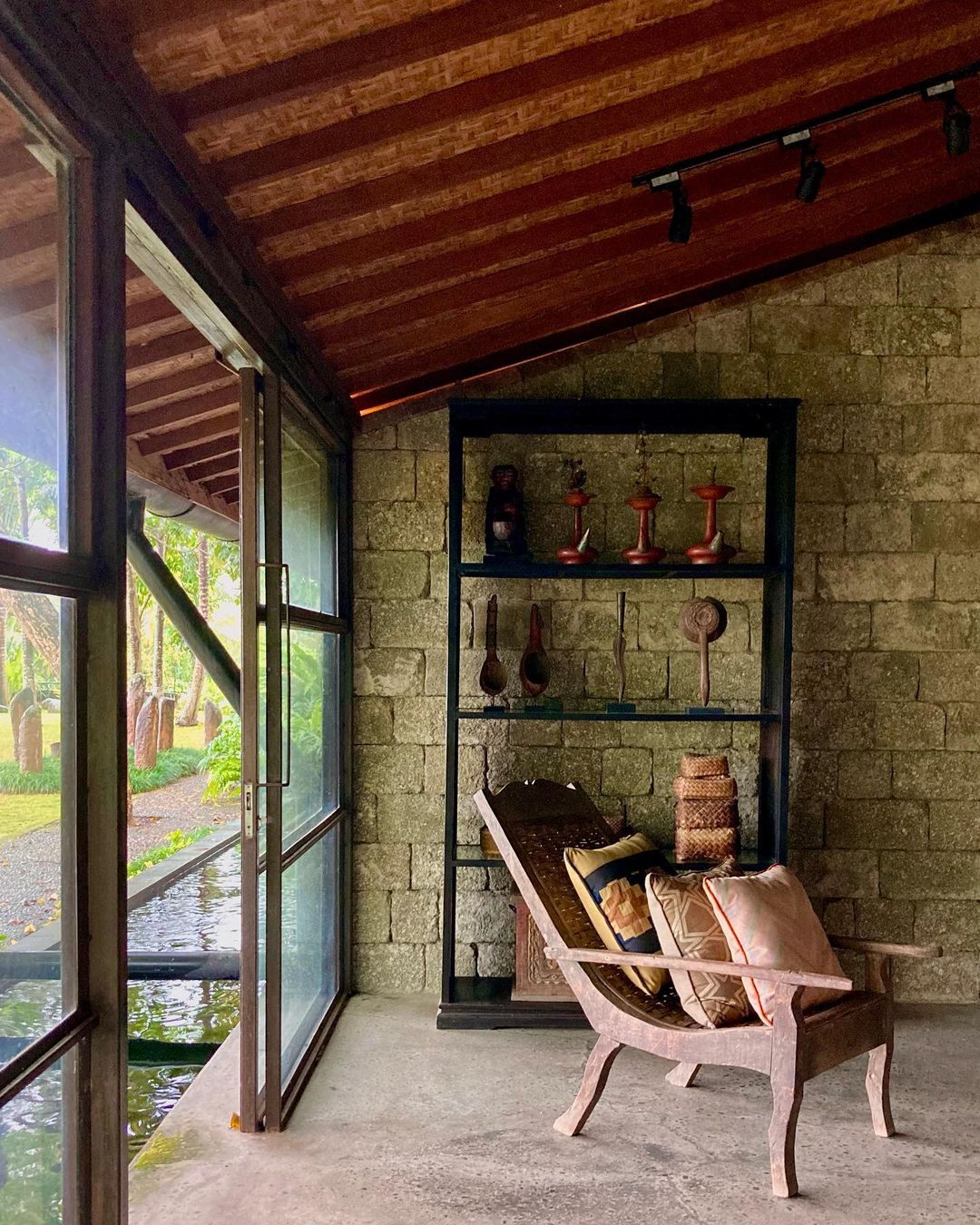 Indoor architectural shot of a wood chaise lounge chair with pillows, open shelving, and stone wall with natural lighting from open doors.