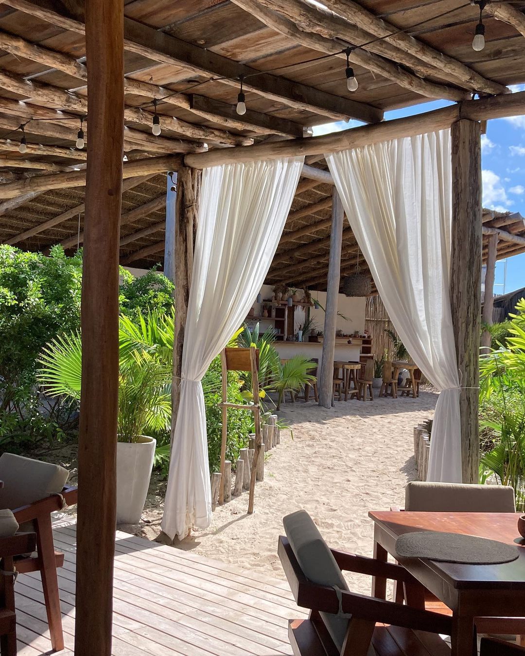 Outdoor restaurant setting with pergola, white curtains, beach sand, and wooden furniture, capturing a tranquil ambiance on a sunny day.