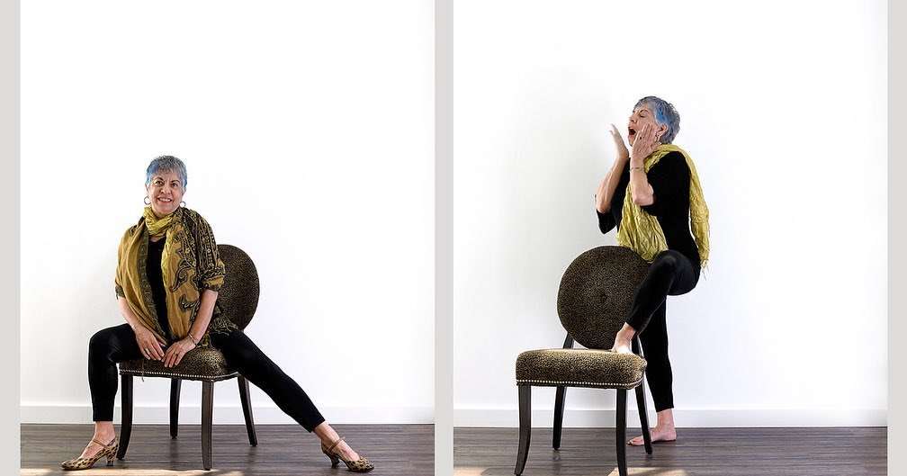Mature woman with short blue hair sitting and standing next to chair in a modern interior; wearing patterned scarf, top, and pants.