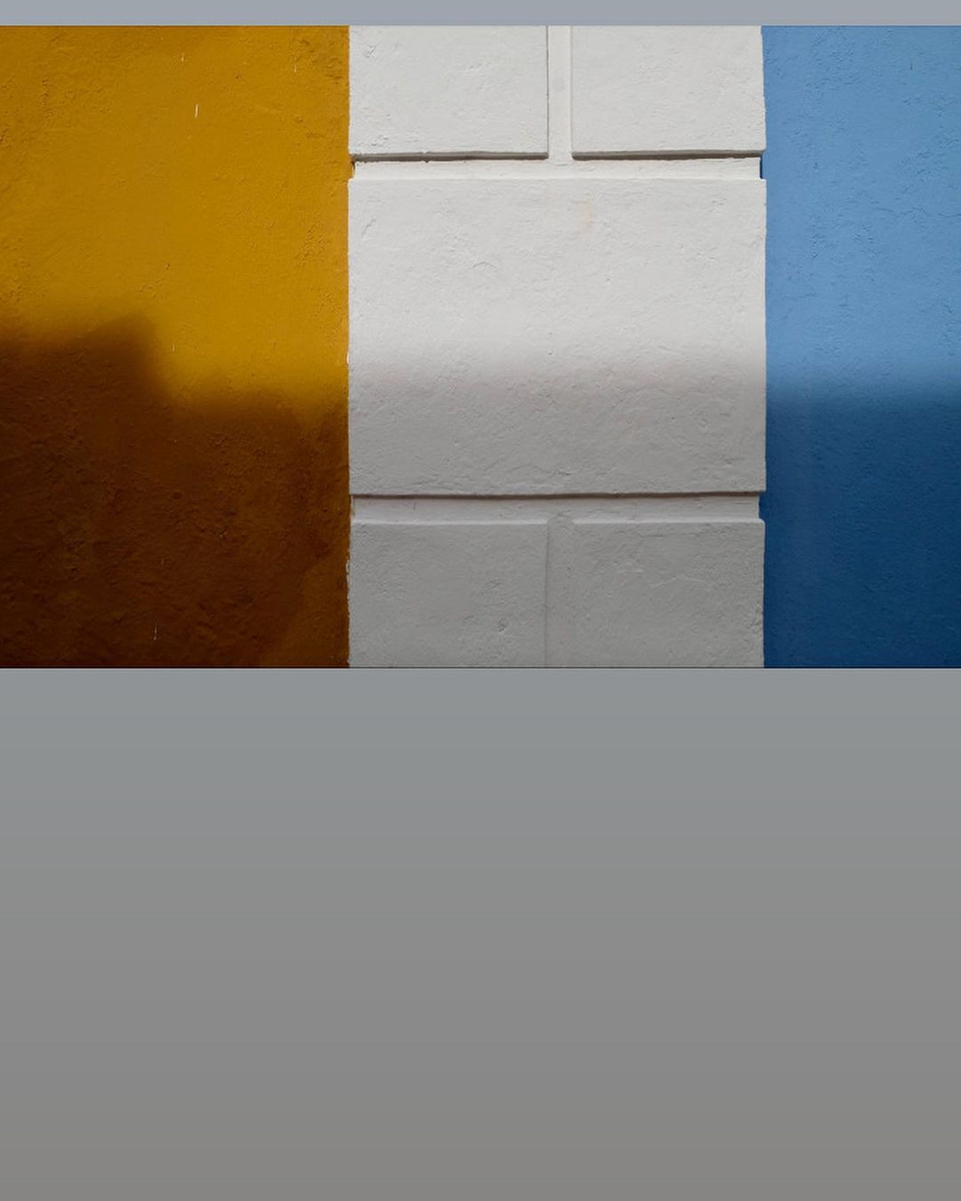 Abstract image of a concrete wall painted in blue, white, and yellow, divided by straight vertical and horizontal lines and sunlight.