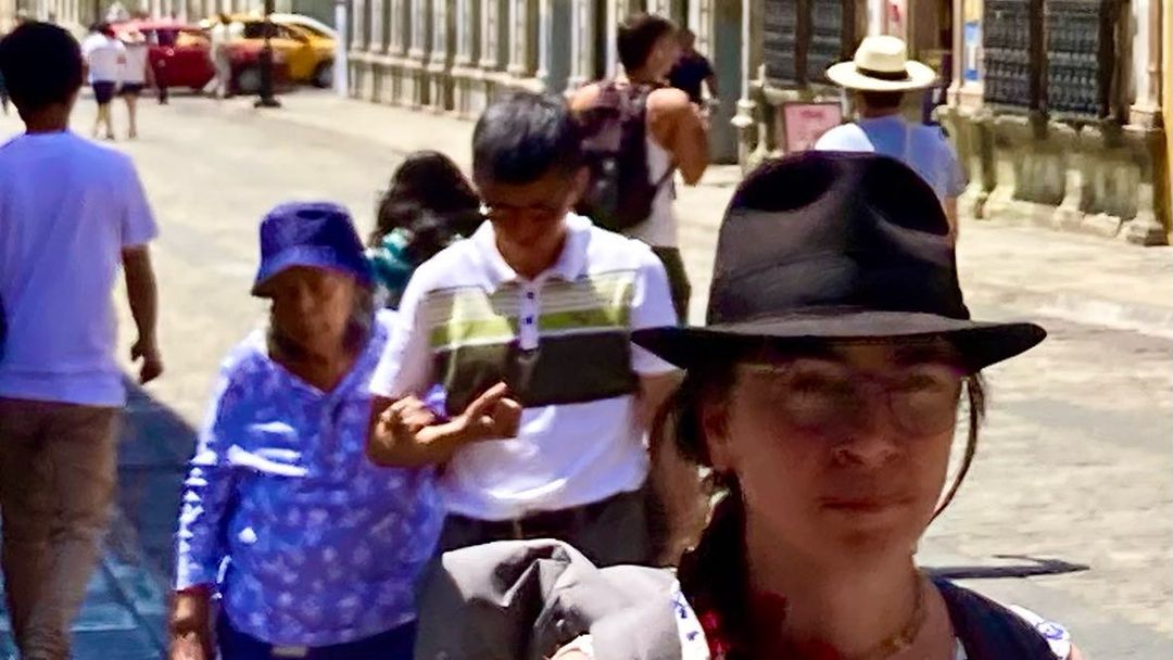 Street scene with a woman in a black fedora hat in the foreground, a small group of pedestrians walking down the sidewalk in the background.