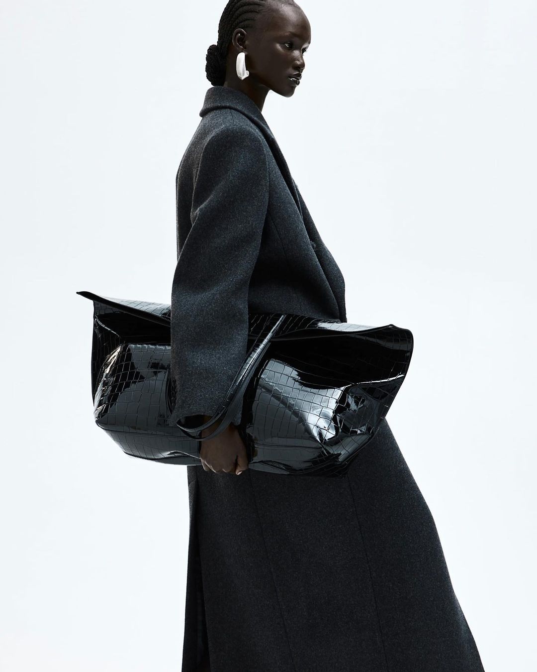 Black woman wearing a dark grey wool coat and holding a large black patent leather handbag against a white background