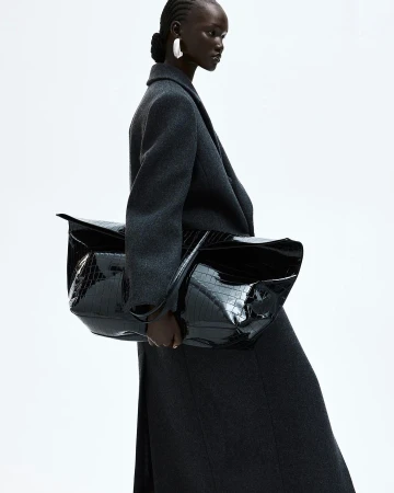 Black woman wearing a dark grey wool coat and holding a large black patent leather handbag against a white background