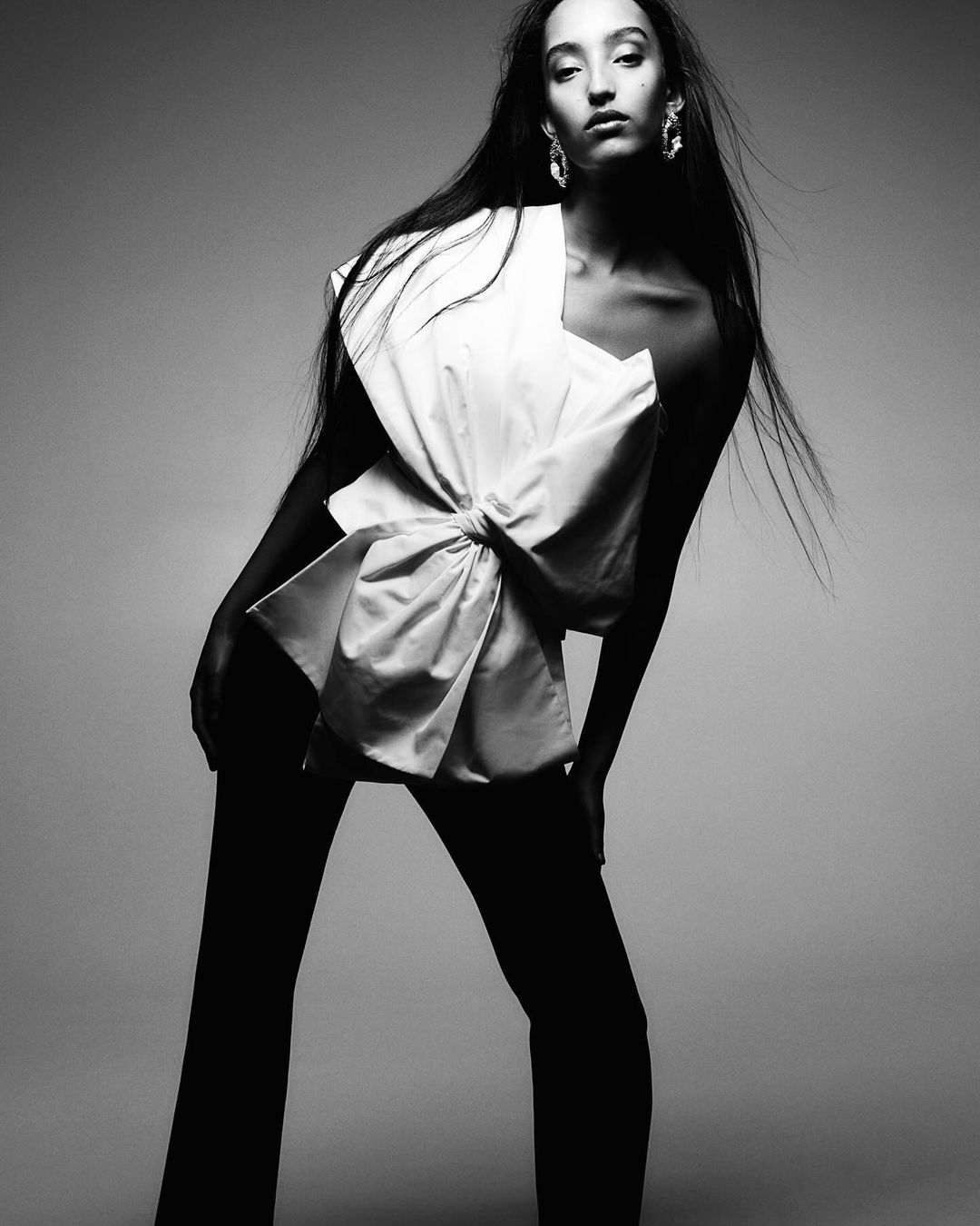 Monochrome studio fashion shot of a young adult female model in a modern asymmetrical top and black trousers, posing confidently.