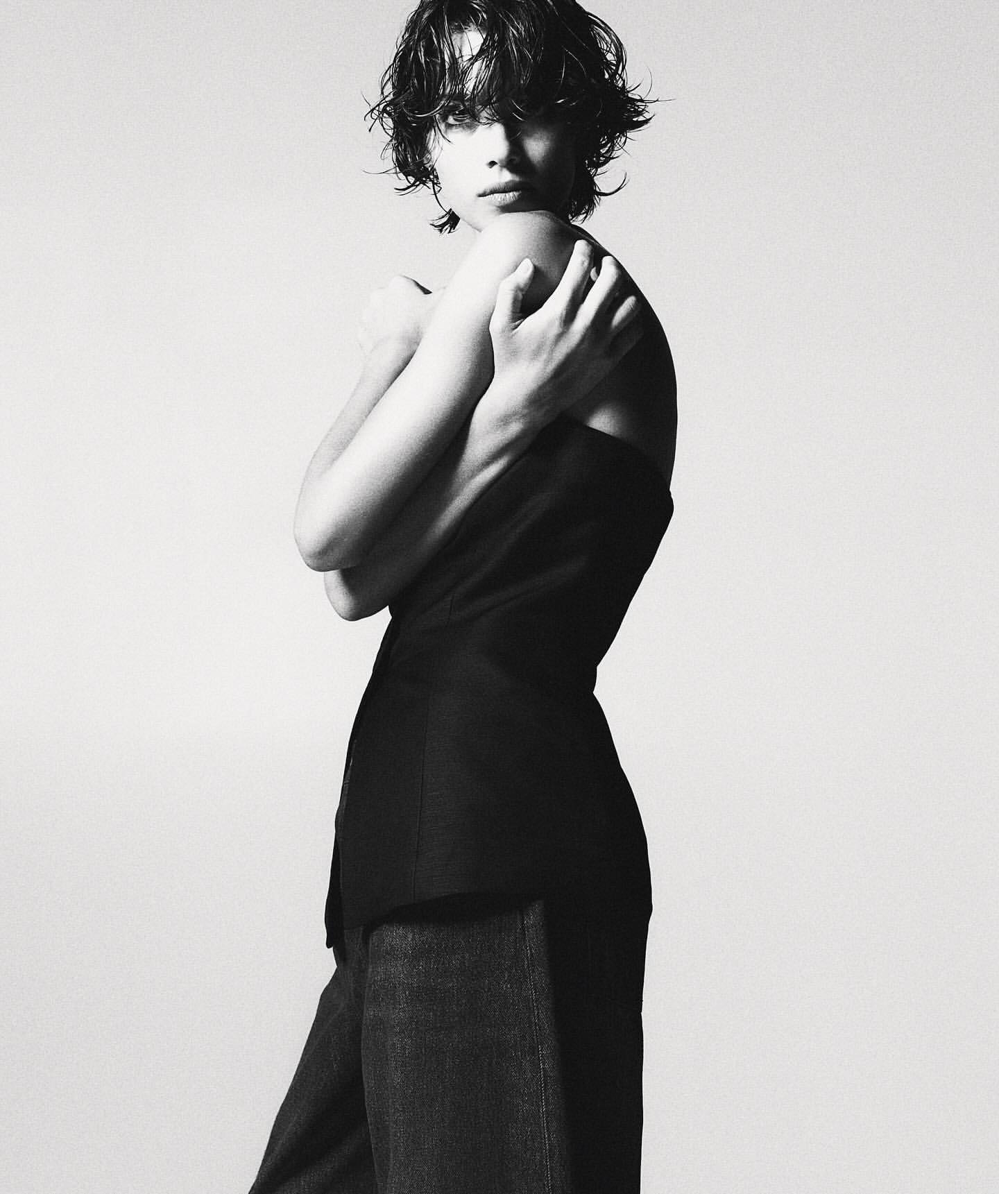 Monochromatic fashion shot of a female model in a dark top and wide-legged trousers, posing indoors against a light background in a studio.
