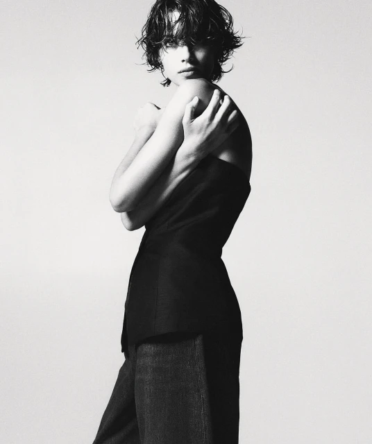 Monochromatic fashion shot of a female model in a dark top and wide-legged trousers, posing indoors against a light background in a studio.