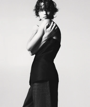 Monochromatic fashion shot of a female model in a dark top and wide-legged trousers, posing indoors against a light background in a studio.