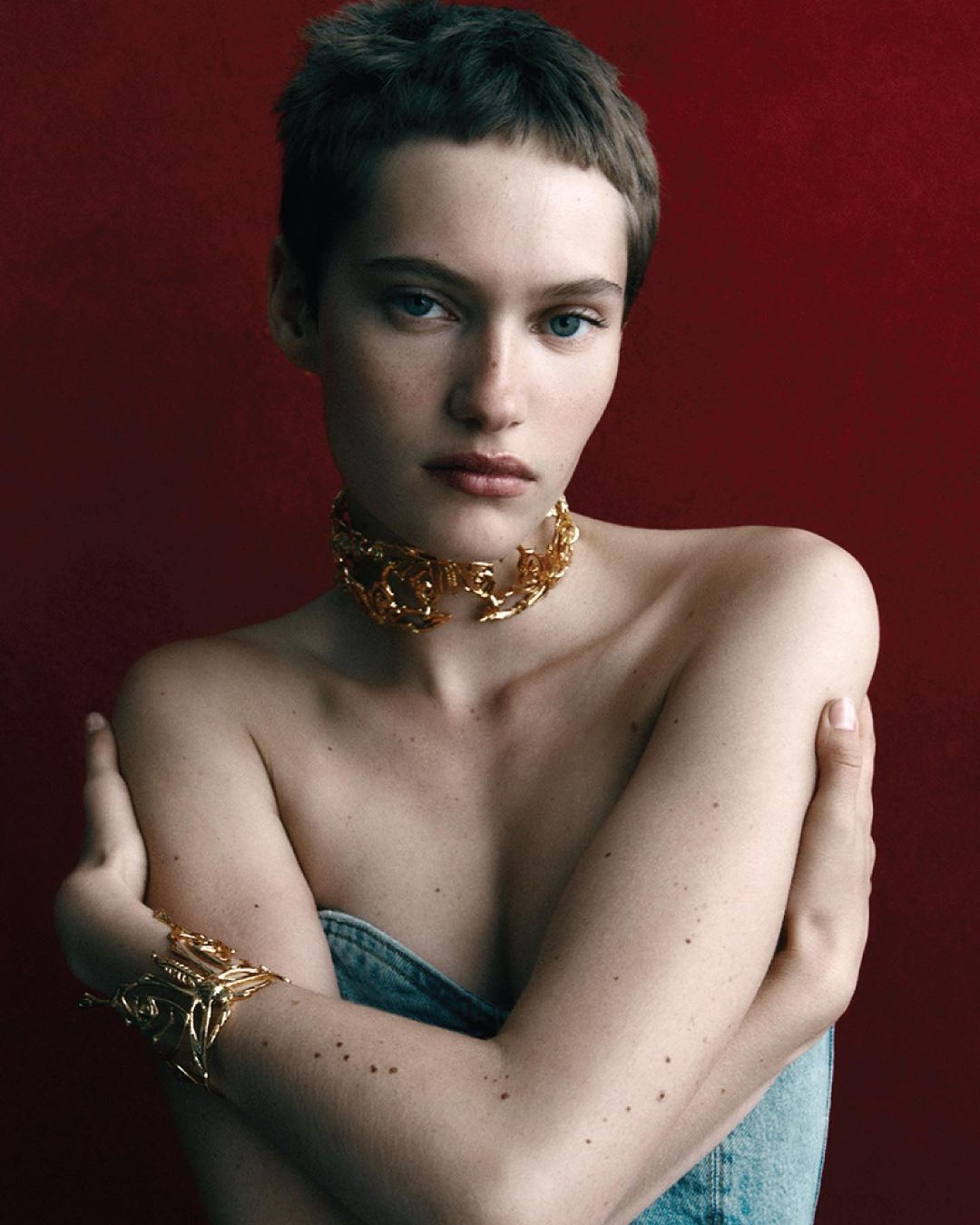 Close-up studio shot of a young, fair-skinned female model wearing a gold choker necklace and bracelet against a deep red backdrop.