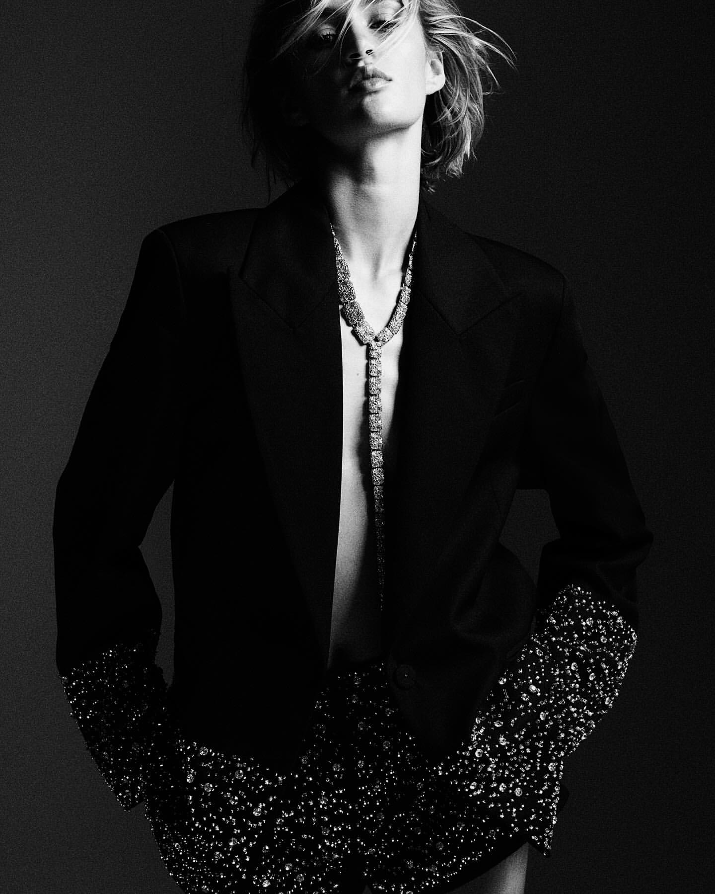 Monochrome portrait of an adult woman in an open blazer with jeweled accents, wearing a long necklace against a dark background.