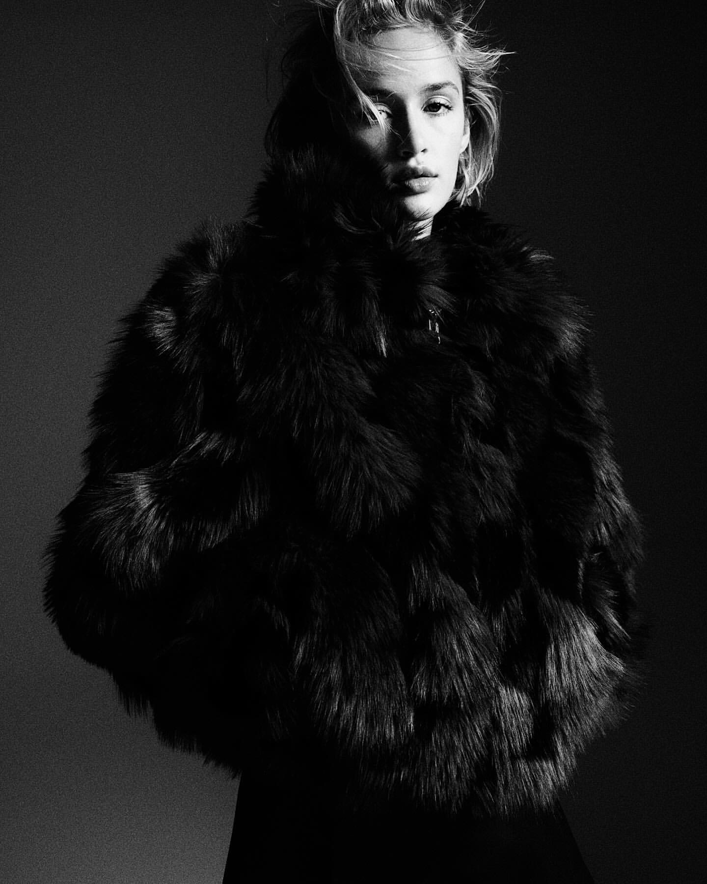 Black and white portrait of an adult female model in a fur coat with messy hair in a studio setting, looking directly at the camera.