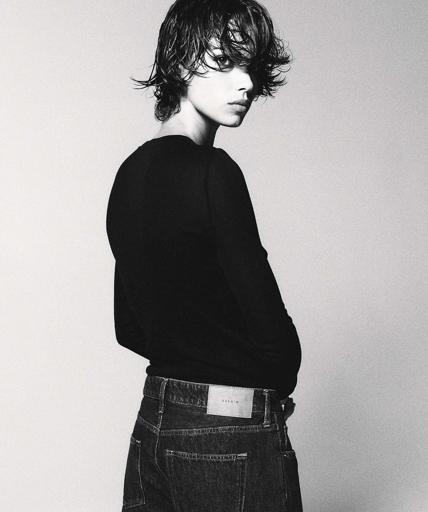 Monochrome studio portrait of a young adult with wet, dark hair, wearing a black top and jeans, posing against a neutral backdrop.