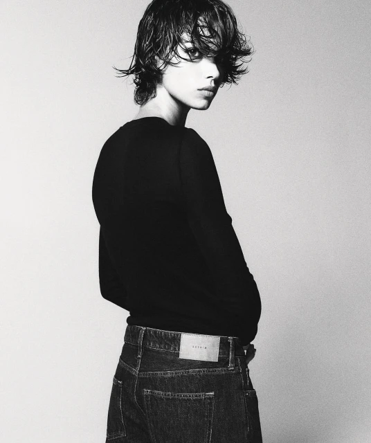 Monochrome studio portrait of a young adult with wet, dark hair, wearing a black top and jeans, posing against a neutral backdrop.