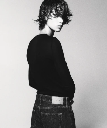 Monochrome studio portrait of a young adult with wet, dark hair, wearing a black top and jeans, posing against a neutral backdrop.