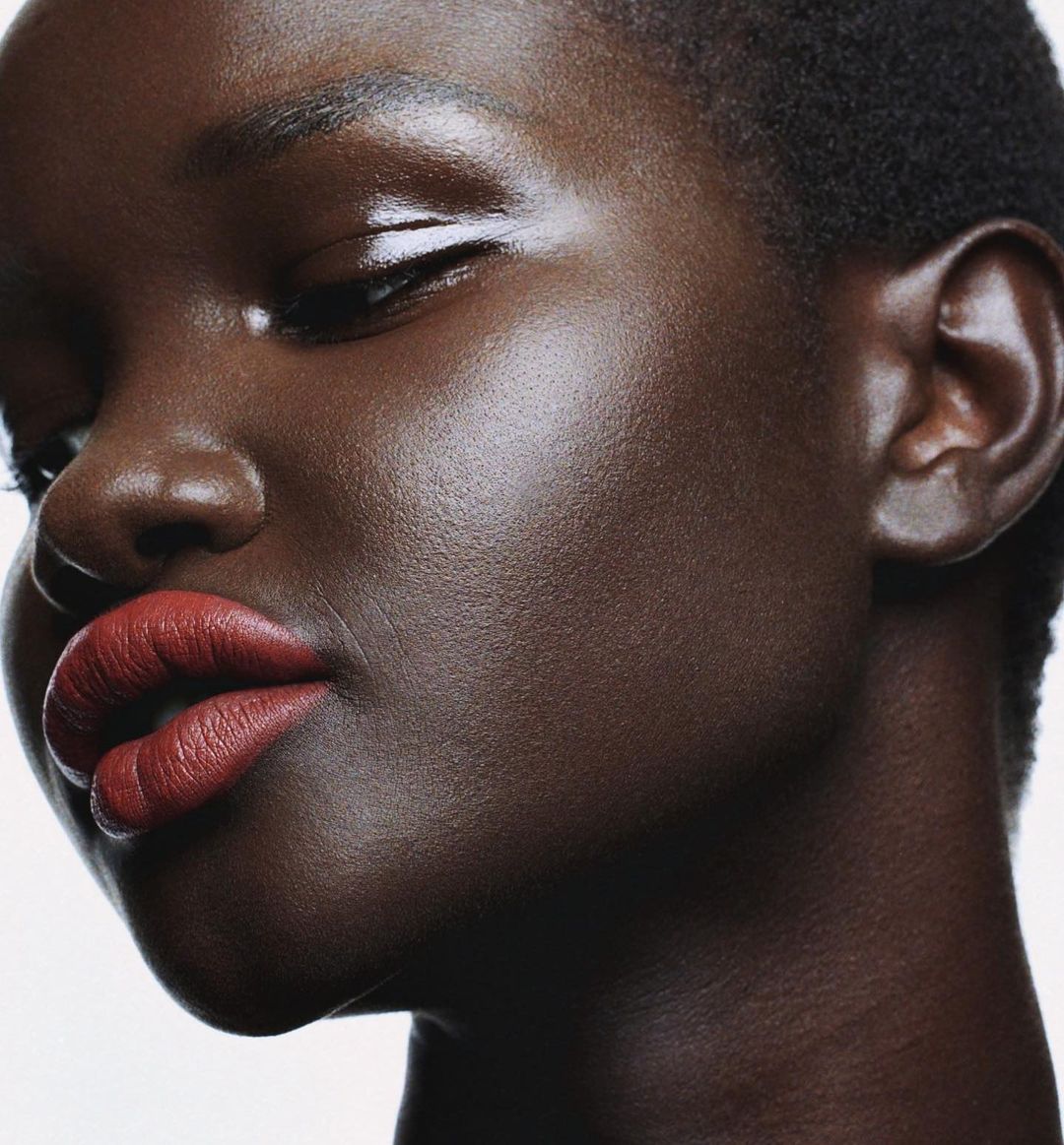 Close-up portrait of a woman with dark skin wearing red lipstick. Her skin is smooth and reflective. Against a white background.