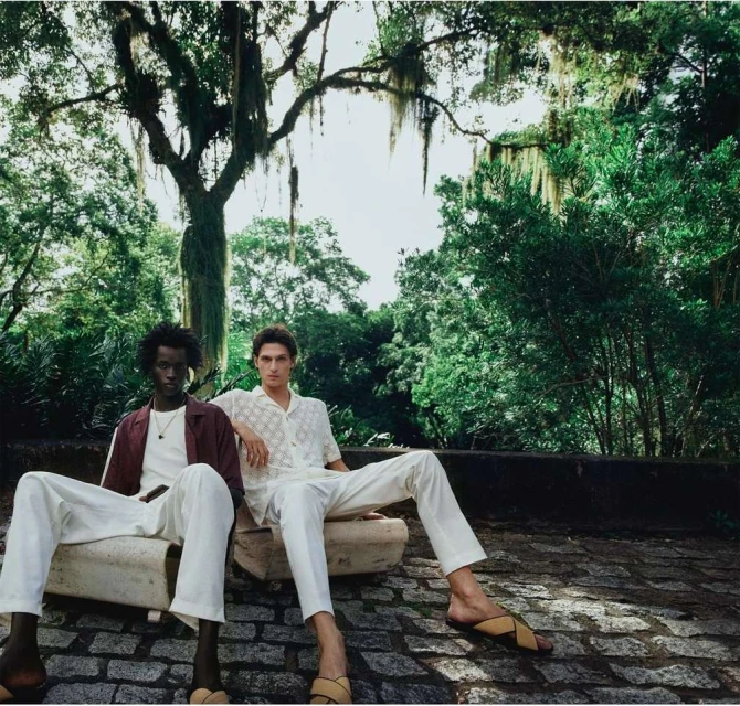 Two men sit outside on concrete structures in casual clothing, surrounded by lush green trees and foliage in a natural setting.