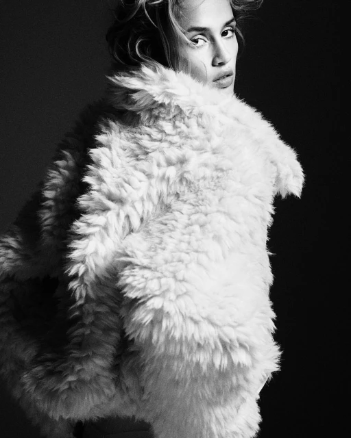 Black and white portrait of a woman with curly hair wearing a large, fluffy white coat, looking over her shoulder.