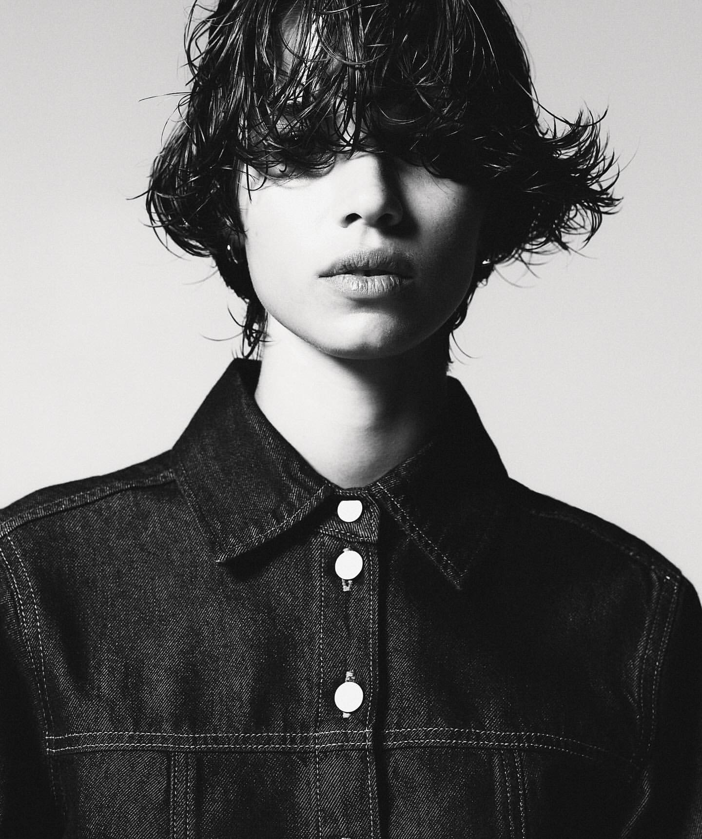 Monochrome portrait of a young adult with wet, dark hair, wearing a dark denim jacket, posing against a light neutral background.