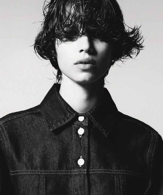 Monochrome portrait of a young adult with wet, dark hair, wearing a dark denim jacket, posing against a light neutral background.