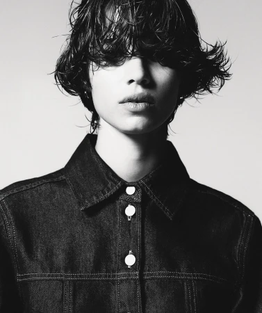 Monochrome portrait of a young adult with wet, dark hair, wearing a dark denim jacket, posing against a light neutral background.