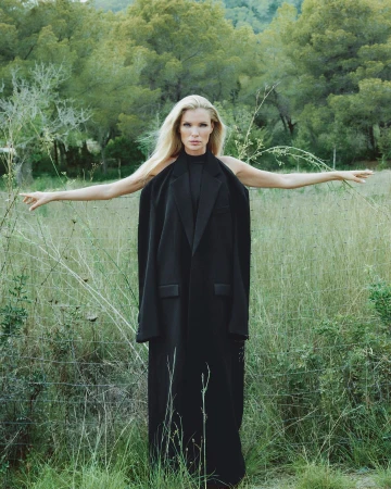 Blonde female model wearing a long black coat standing in tall grass outdoors with arms outstretched.