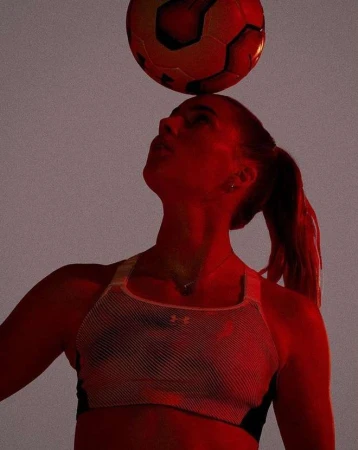 Adult female balances soccer ball on head in a medium shot with red lighting; wearing a sports bra in a gray indoor setting.
