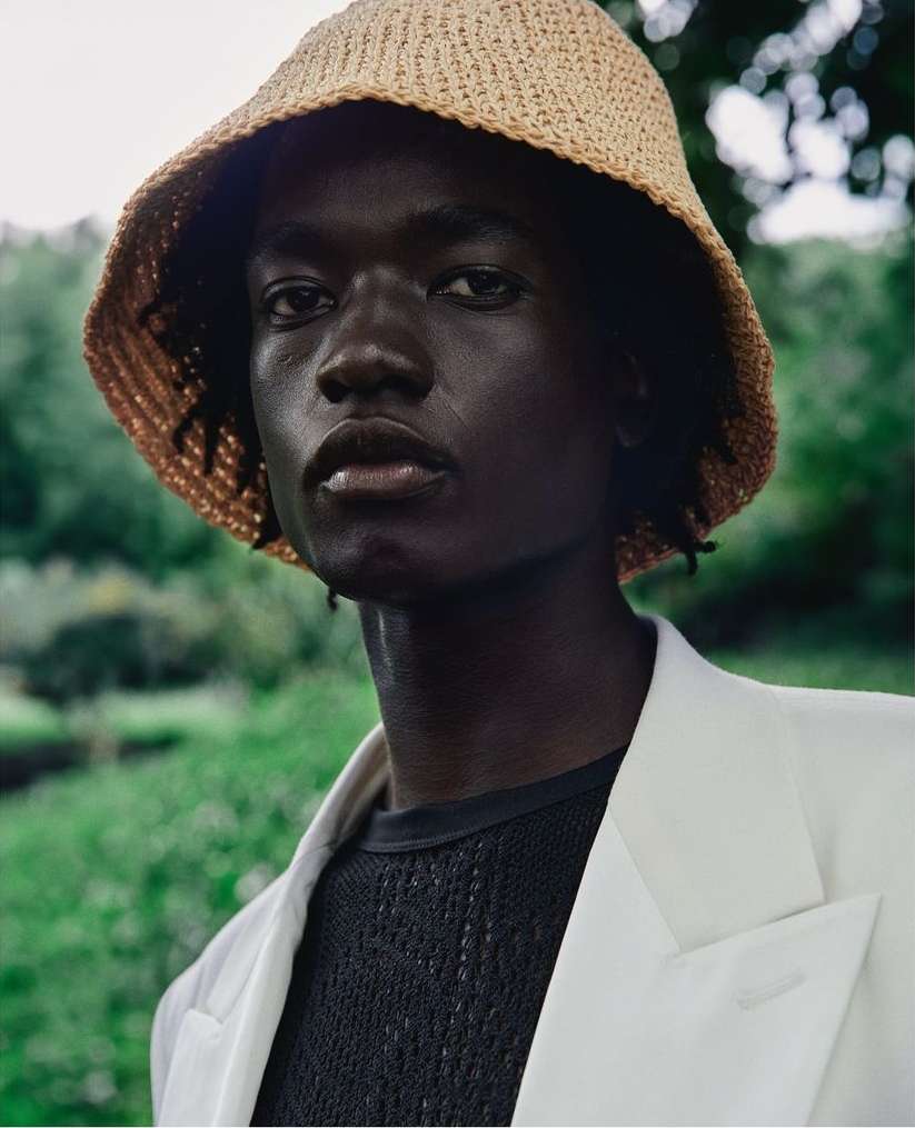 Portrait of a Black man wearing a straw bucket hat and white blazer outdoors by Carlos Garcia
